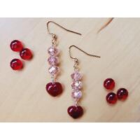 Light Pink Beaded Dangle Earrings with Small Red Heart Charm lays on a table surrounded by red beads. Gold plated fish hook ear wires.  Light Pink Beaded Dangle Earrings with Small Red Heart Charm lays on a table surrounded by red beads. Gold plated fish hook ear wires.